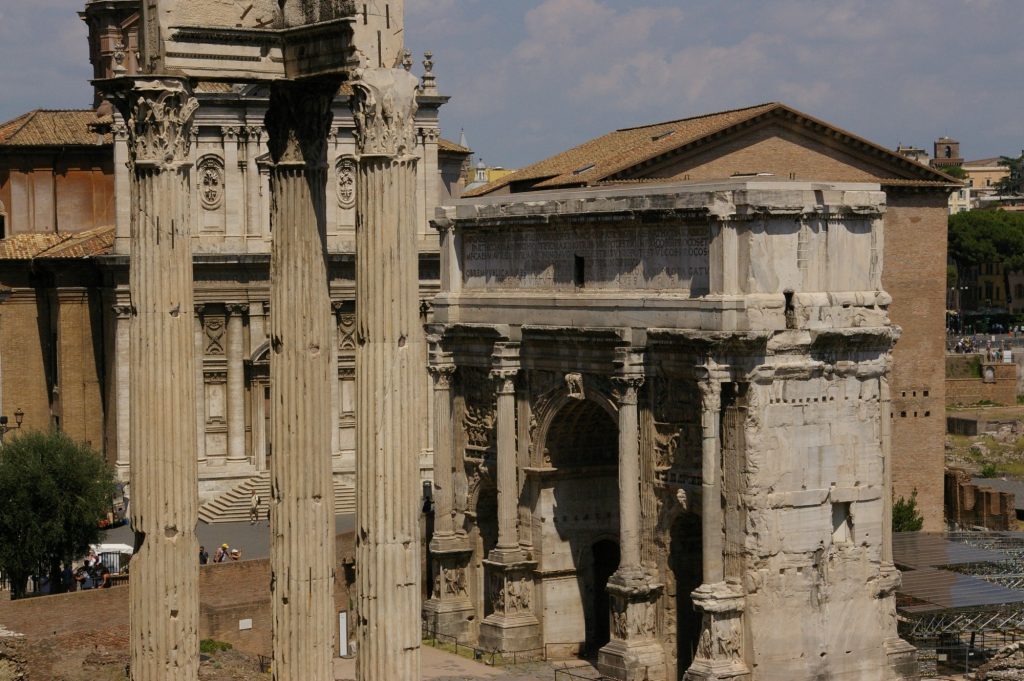 In this view, 2 elements of the forum are visible. One is a set of 3 very tall corinthian columns holding up the remains of a pediment. The other is an archway with columns in front and the entire roof/pediment still intact.