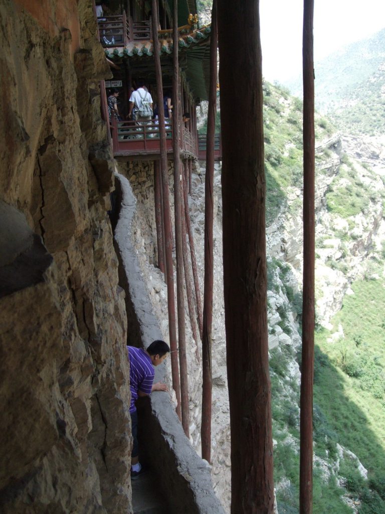 A narrow walkway along the cliff, with long support beams supporting the monastery above.