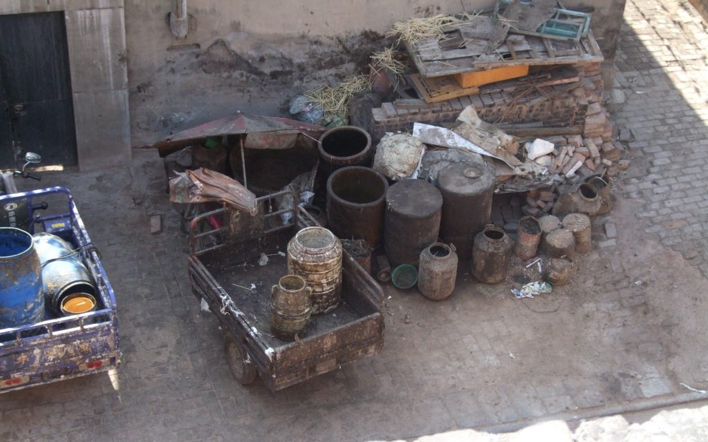 Seen from above: Two wagons are parked, with barrels on them of various sizes, all looking brown and filthy. Beside them is a cluster of more barrels, again in a range of sizes, some open, some closed. the ground is paved in brick, but damaged in places.