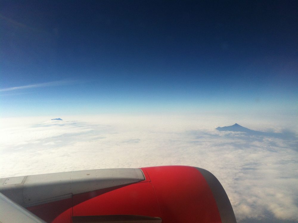 view from a plane looks over a red-painted engine and a cloud-covered land, except for one mountain in the distance that peeks out through the cloud.