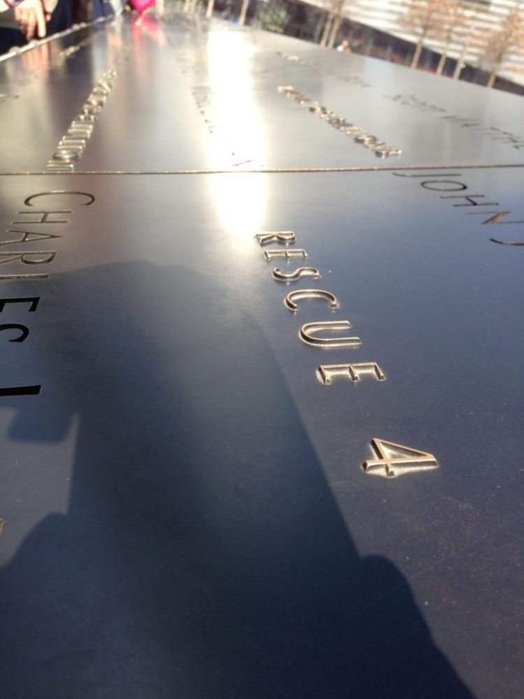 a detail of the 9/11 memorial in New York City: a dark shiny marble wall with raised letters spelling out the victims' names and their affiliations. 