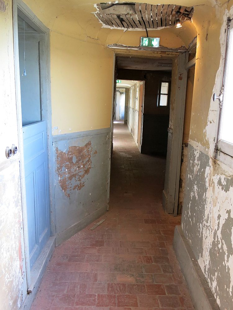 The hallway is in very poor condition: the walls are painted blue below and yellow above, but the paint is badly peeled and on one side the lower part of the wall - the wainscoting, has come off entirely. A patch on the ceiling is missing its plasterwork so the slats of wood are visible.