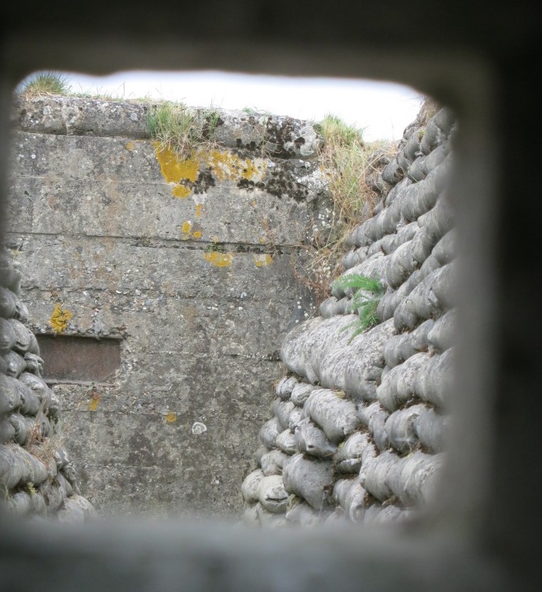 Looking through a peephole down a trench. The trench is sided with piled-up sandbags (actually concrete filled in this restored trench). The trench ends at a rough concrete wall that also has a small peephole in it.