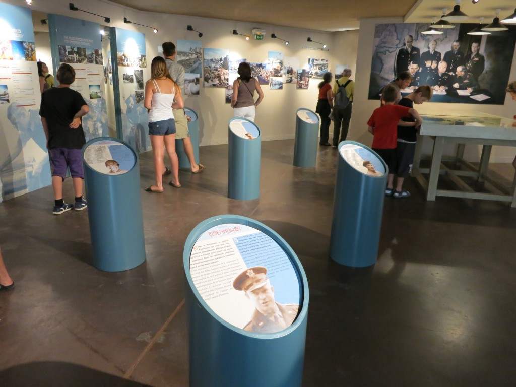 On the far wall: lots of texts, photos and maps. Several people look at the wall, their backs to the camera. The space between them and the camera has 5 pillars that go to about waist-height, each one with a flat angled top. Each has a photo center bottom of the column's top, with text above the photo. On the nearest one it is possible to read the title at the top, which says Eisenhower.