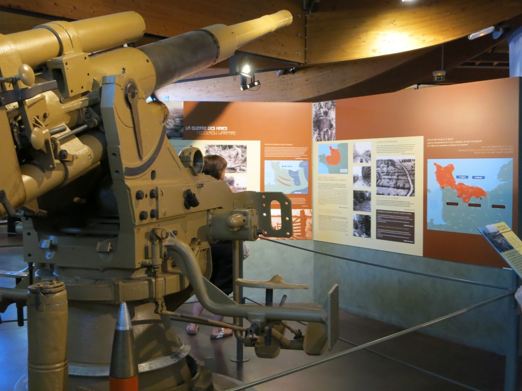 Left to center of the image is a large artillery piece: a cannon? a machine gun? Behind it is a wall with an assortment of maps and photographs on it, accompanied by blocks of text.