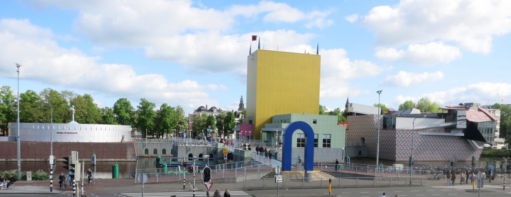 An upside-down U forms the entrance. Beyond that, a bridge, then parts of the museum on the left and right. On the right, a tall yellow building with no windows. Also a smaller light green building. Further right, a building that is very oddly shapped, with part of it sticking out at an angle. On the left of the bridge is a low, round, grey building. 