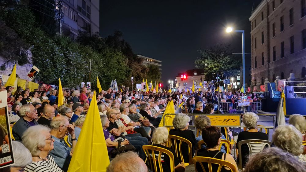A crowd of people standing or sitting on chairs looking at something outside of the picture on the right.