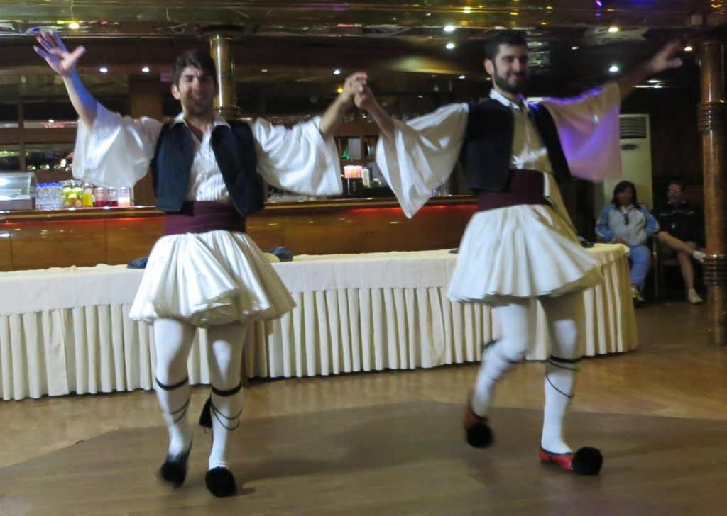 two men dancing, arms raised, wearing white skirts, and big-sleeved shirts and a black vest.