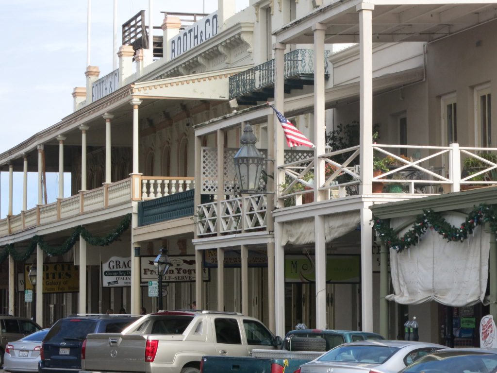 Buildins are two or three stories tall. All are painted a beige color and have balconies held up with poles, creating a shaded sidewalk. 