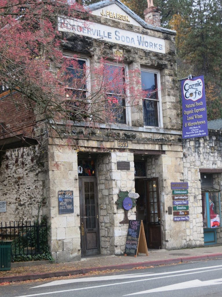 A two story stone building. Reading top to bottom, signs say J. Pearson, Placerville Soda Works, Cozmic Cafe Natural Foods Organic Espresso Local Wines & Microbrews. 