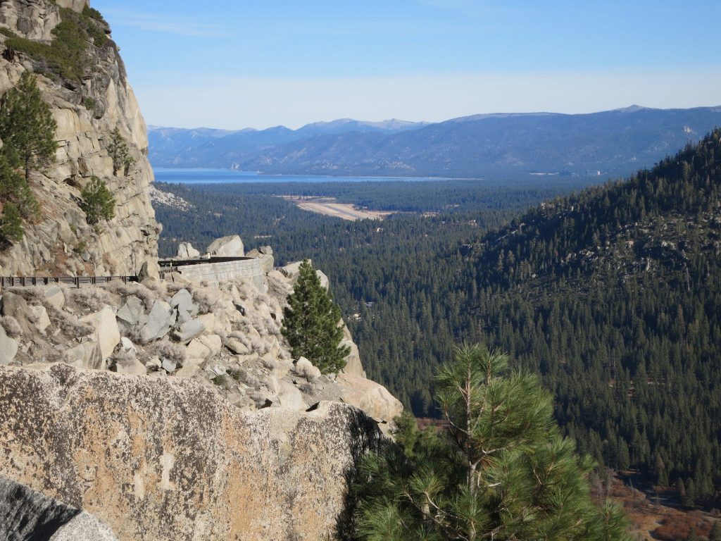 On the left, a bare rock cliff, with a road cut along it, with guard rails on the cliff side. Ahead, a tree-filled valley. Beyond that, a bit of the lake is visible and mountains beyond that.