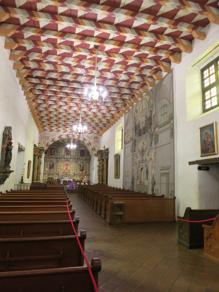 view of the interior of Mission Dolores with painted ceiling: a long narrow room, looking toward the altar in the distance, which appears to have a number of figures painted on its back wall.  An aisle down the center of the room with pews on either side. The ceiling has heavy beams going across and they and the spaces between them are painted with a simple geometrical pattern of rectangles and triangles in dark red, white, grey and mustard yellow. ON the right-hand wall is a large mural, but it's hard to see what it depicts from this angle.