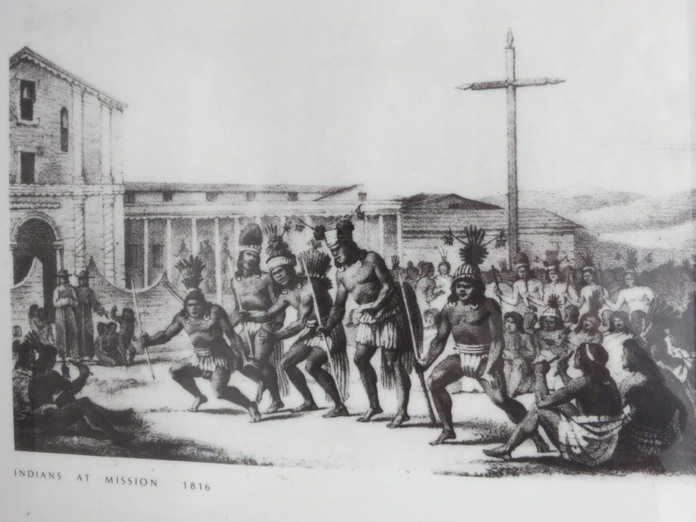 drawing of Native Americans at Mission Dolore: Black ink on paper, says "Indians at Mission 1816" at the bottom. 5 Native American men wearing just clothes around their bottoms and headdresses stand in a row holding spears. They appear to be dancing. Behind them many more Native Americans sit on the ground, watching them. 