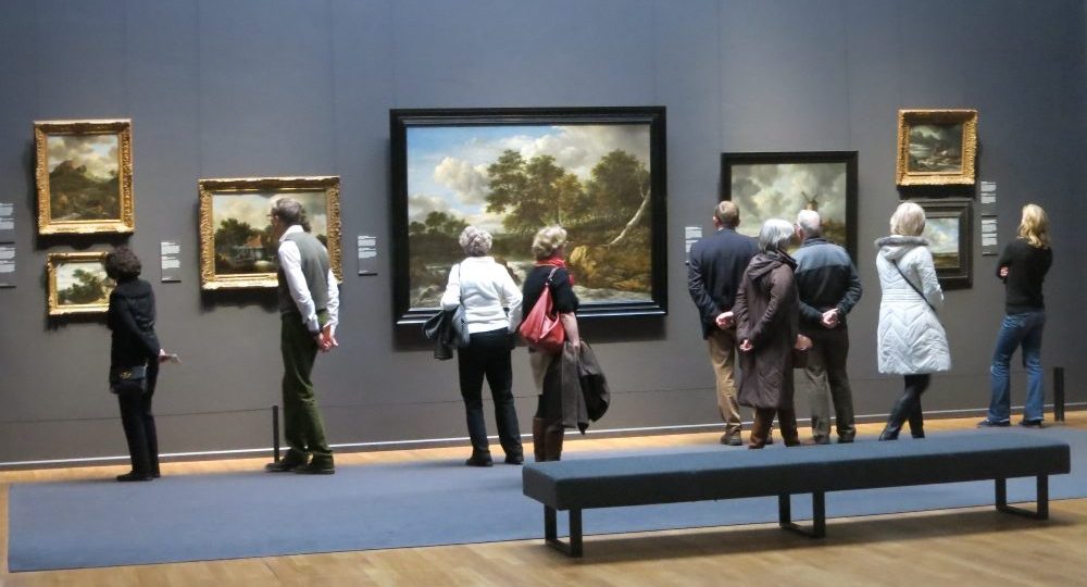 A row of Golden Age landscapes on the wall, and 9 visitors, backs to the camera, viewing the paintings.