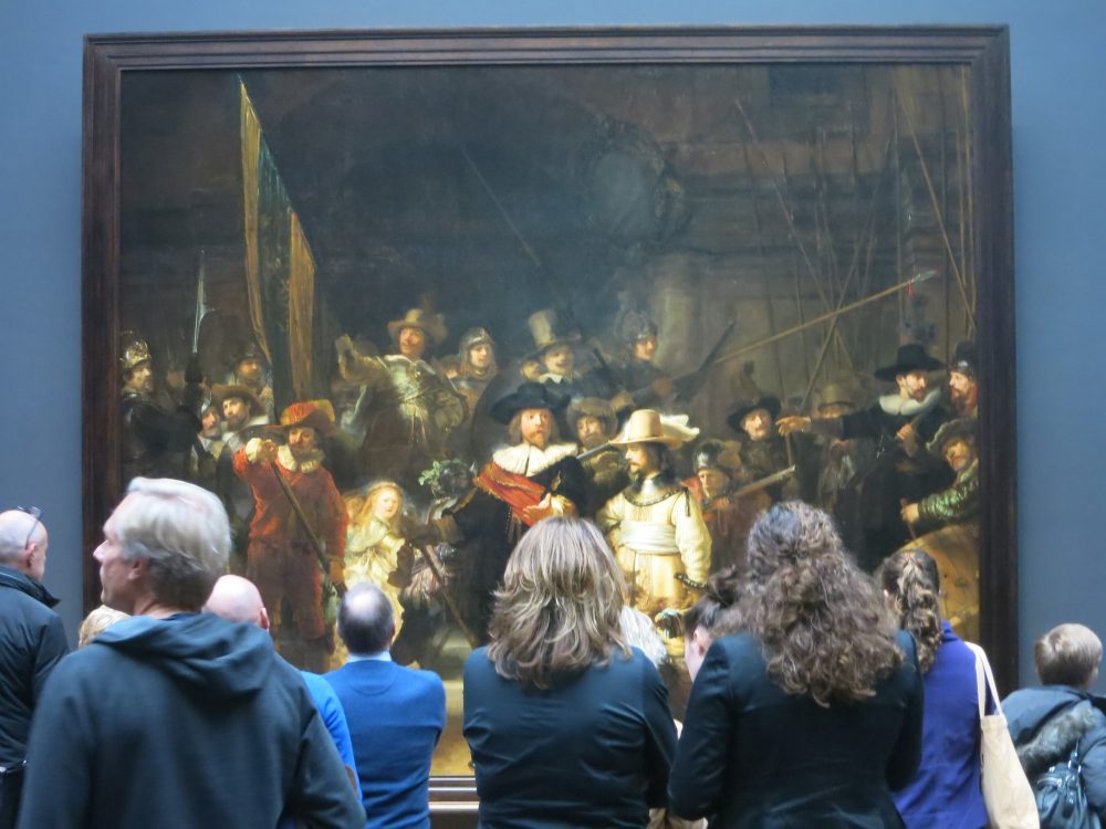 The Night Watch is a large painting of a group of men in the 16th century. In front of the painting, a group of people, backs to the camera, obscure the bottom half of the painting. They are all coincidentally wearing blue jackets, and the wall around the painting is also blue.