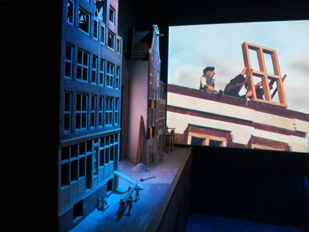 On the left are blue-lit model houses and on the far wall a piece of a film is visible showing workers installing a window frame. Canal House Museum Amsterdam