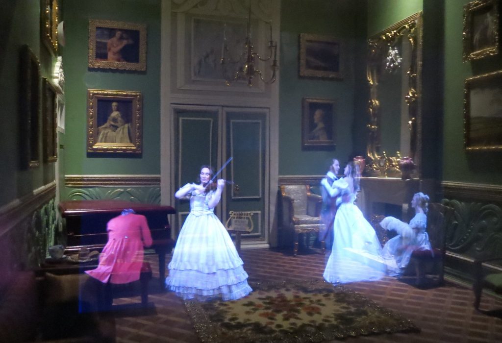 In an elegant room lined with portraits, a woman plays guitar in a long white dress, a man in a red coat plays piano, and several other people dance.