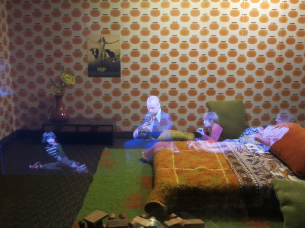 Sitting on the floor, a man in a 60's era room plays guitar for his children. At the Canal House Museum in Amsterdam