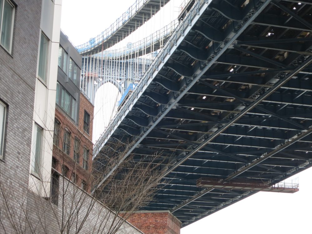 view of the Manhattan Bridge from underneath: It extends from the bottom middle of the photo to the top right-hand corner. On the left a bit of a row of buildings is visible. New York City on a Budget