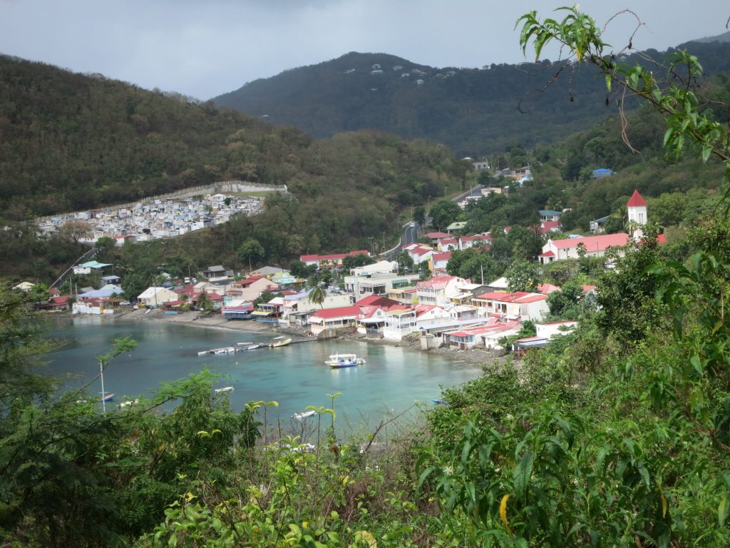 This is the same view of Deshaies that is used in the opening credits of "Death in Paradise", except with a wider view so that the cemetery on the side of the hill beyond the village is also visible. In this photo, it looks like a small town with small houses encircled by a wall. Seen from a hill above the village, the bay is in the center bottom of the photo and the buildings cluster around it: mostly white with red roofs. Up the hill from the bay is the church, with its white tower with a red pointed roof. Around and behind the small village is all green growth.