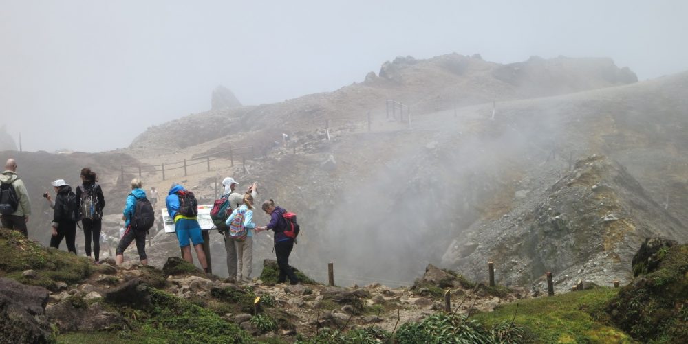 a group of hikers, seen from behind, stand in a row looking at a hole that is partly visible behind them. White smoke is visible rising from the hole. 
