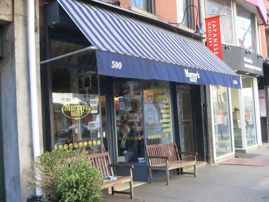 A simple storefront: glass windows with a door between them, two benches in front outside. An awning, striped in blue and white, extends out above the entrance and both benches. It says "500" "Murray's bagels" and "500" along its edge.