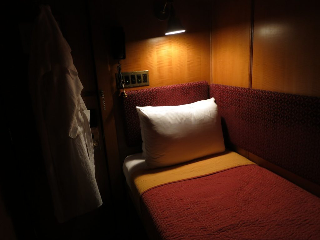 This interior view shows a single bed with a light above the pillow and paneling on the wall.
