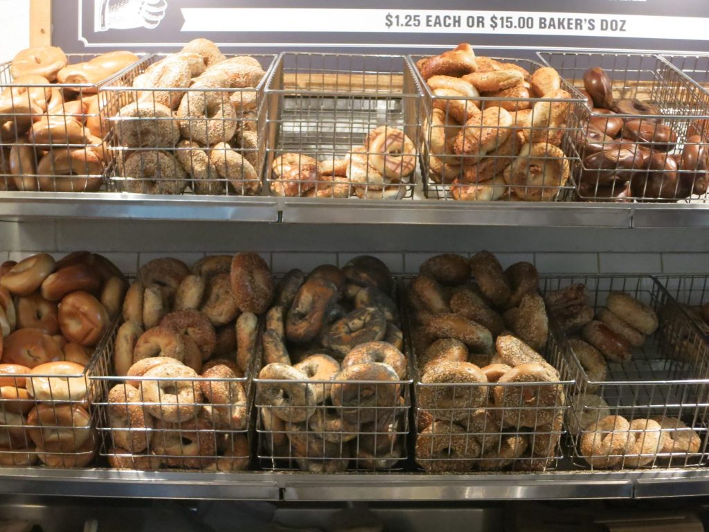 Lots of different types of bagels, displayed in two rows of baskets in a bagel store. "$1.25 each or $15.00 baker's doz"