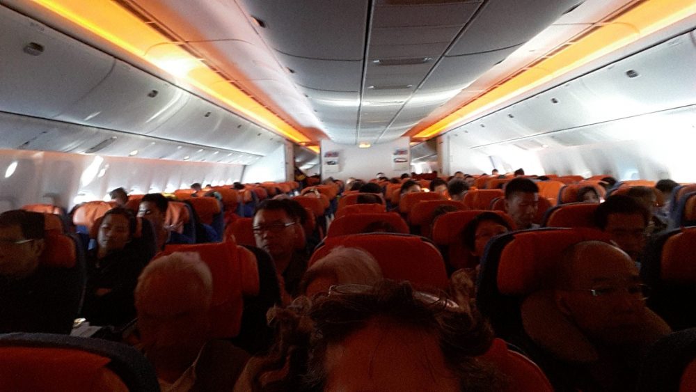 view of the interior of the Aeroflot Boeing aircraft, looking toward the back. The people are seated in red chairs and the lights along the baggage compartments are yellow.