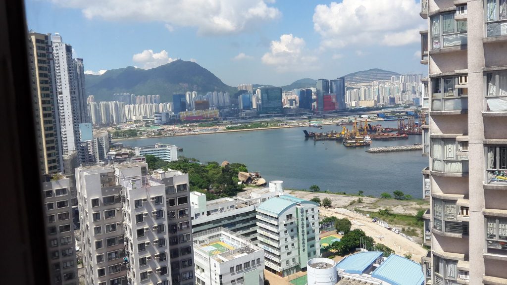 view of a section of Hong Kong from my airbnb apartment across the way in Kowloon. Tall buildings to the right and left. The sea straight ahead with a section of Hong Kong - tall buildings in a cluster in front of forested mountains - beyond the sea.