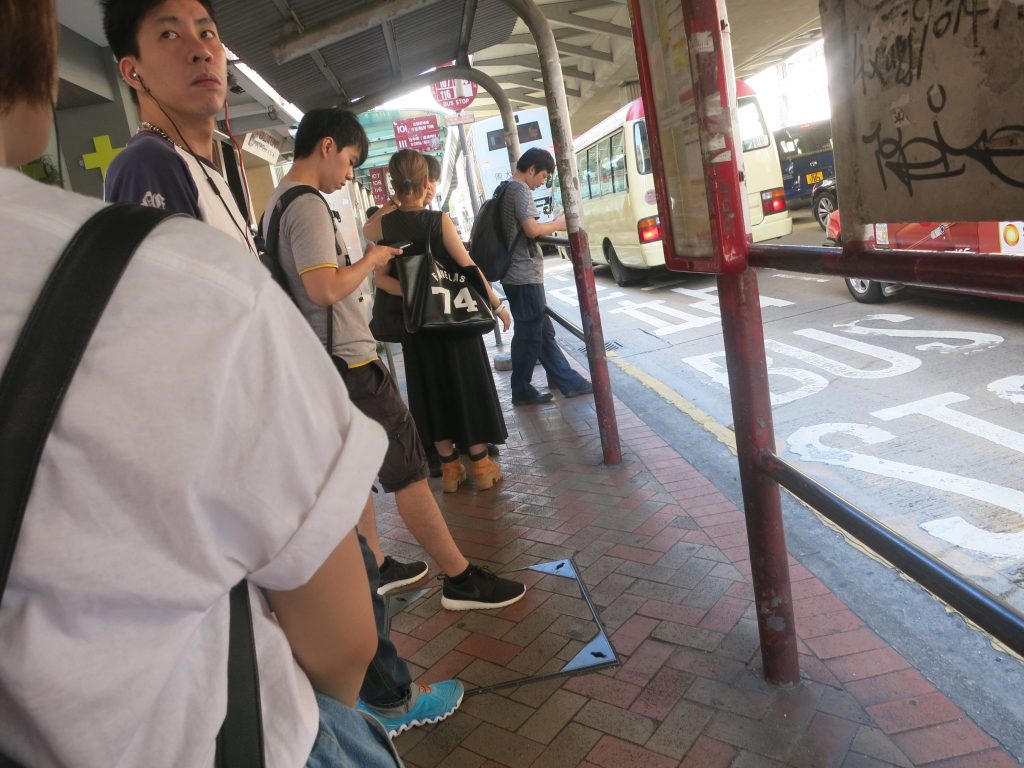 a bus station with people waiting for the bus in Hong Kong: most are looking at their phones. They stand under a simple roof, held up with poles, next to a lane of the street painted with the words "bus stop".