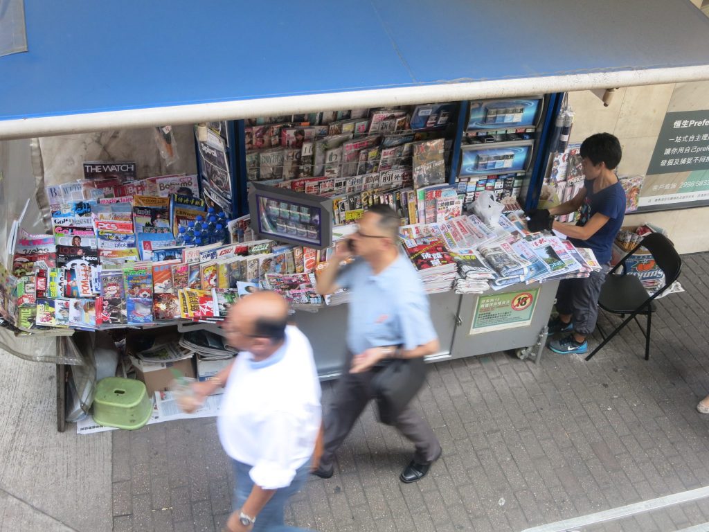 view down from a bus window, shows a newsstand in Hong Kong, with a woman working there, and pedestrians passing. The newstand is covered with lots of magazines and newspapers and has a blue awning above it.