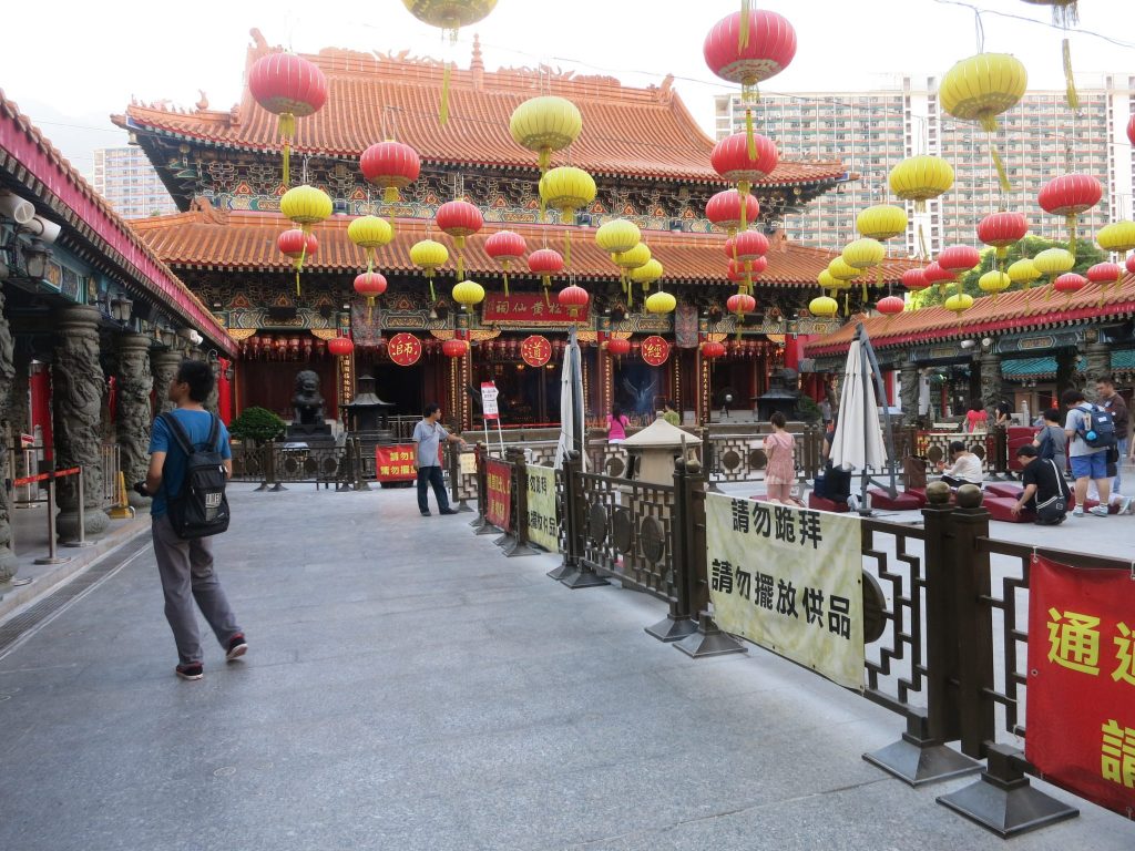 Temple in the background with the traditional roof line that curves upwards at the edges in red tiles. The open space in front has strings of paper lanterns hanging over it in lines of red and yellow. A few people are visible near the temple, praying. Behind the table some tall buildings.