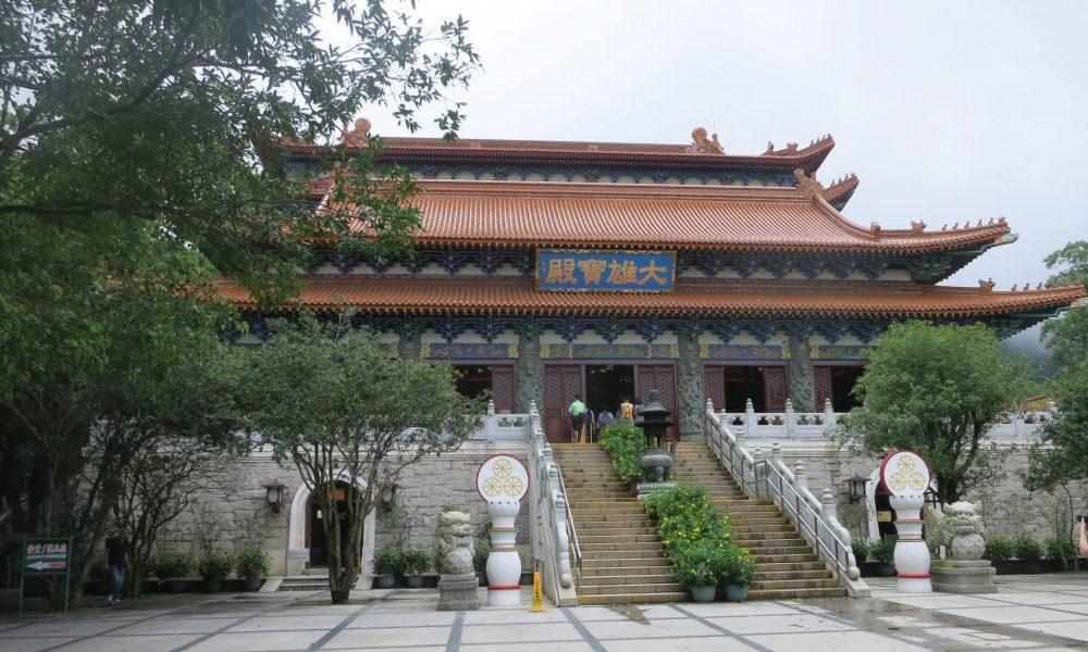 Decorated in a traditional Chinese style, the Po Lin monastery temple contains three Buddha statues. A large stairway leads up to its center. The roof is red and curves in the traditional Chinese style. 