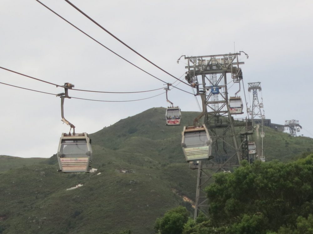 view of the cable car line from the end near the Big Buddha. The wires curve down between each of the supporting towers, with the small cars hanging from the wires: square boxes with windows. The landscape is green and mountainous.