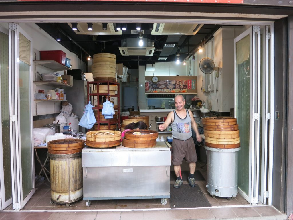 storefront, open to the street in Hong Kong, with large steam baskets holding a variety of dim sum. The owner stands next to where the steam baskets are displayed and smiles for the camera.