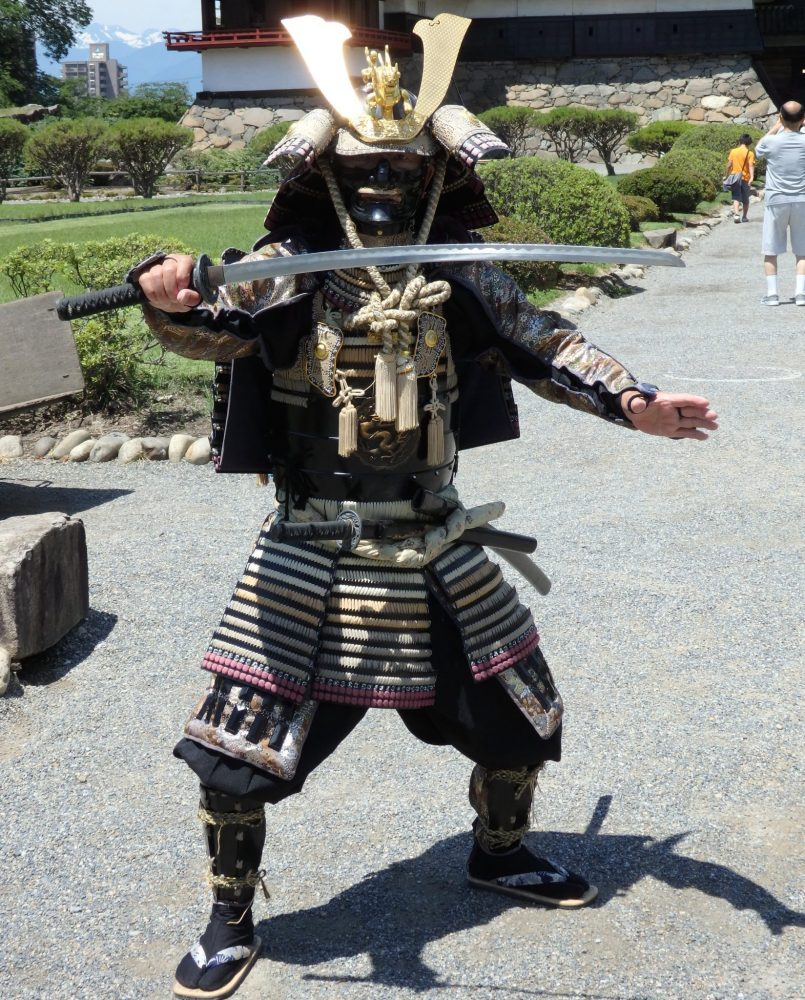 A man in full samurai armor blocks entrance with his sword, in front of Matsumoto Castle.
