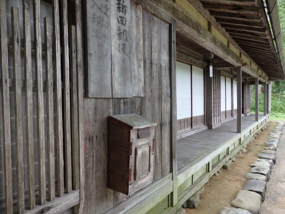 Wooden house, looking along its front. A rather narrow porch shaded by the eaves of the roof.