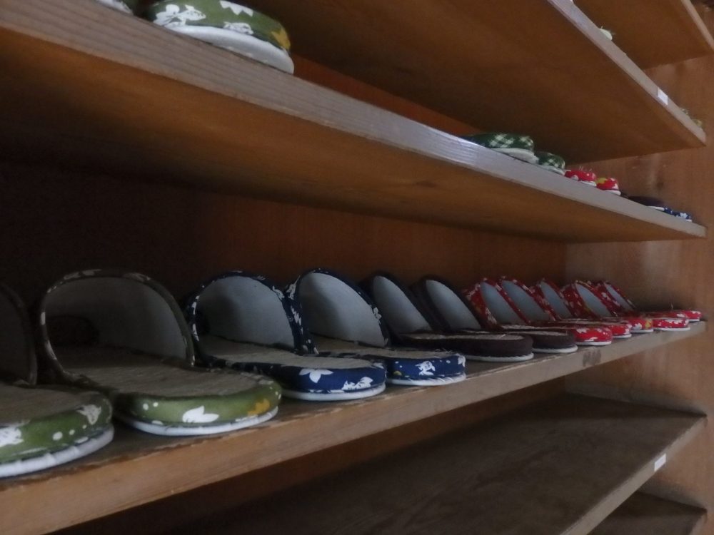 The slippers have a flowered pattern, some in green, some in red and some in blue. They are lined up very neatly on what looks like a bookshelf. 