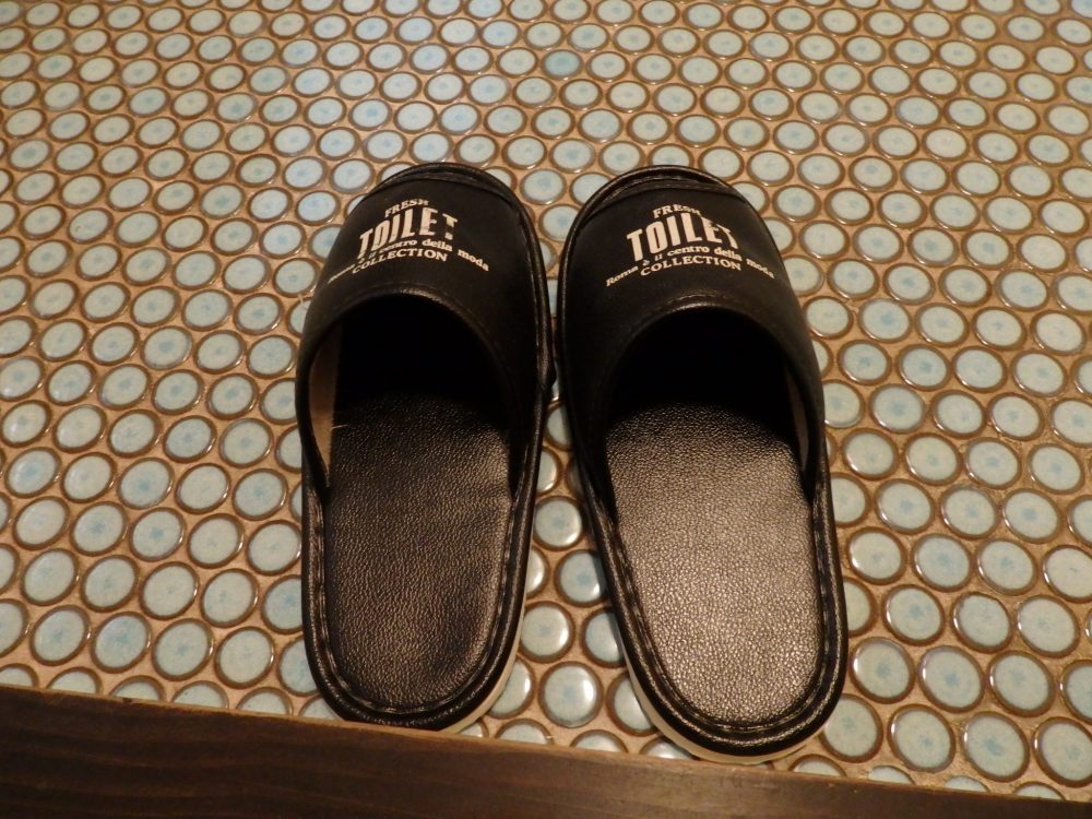 A pair of brown vinyl slippers on a rubber mat. The slippers say TOILET on each one.