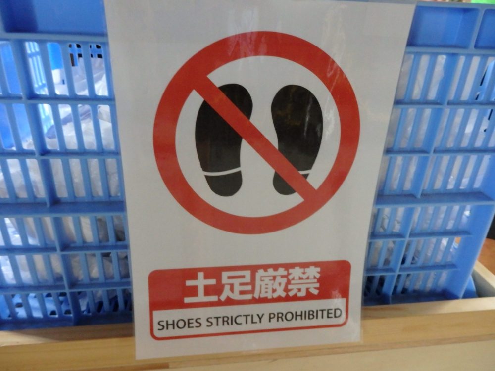 a sign reading "shoes strictly prohibited" in Japanese and English. Above that, a symbol of two footprints with a red circle around them and a red slash across them.  