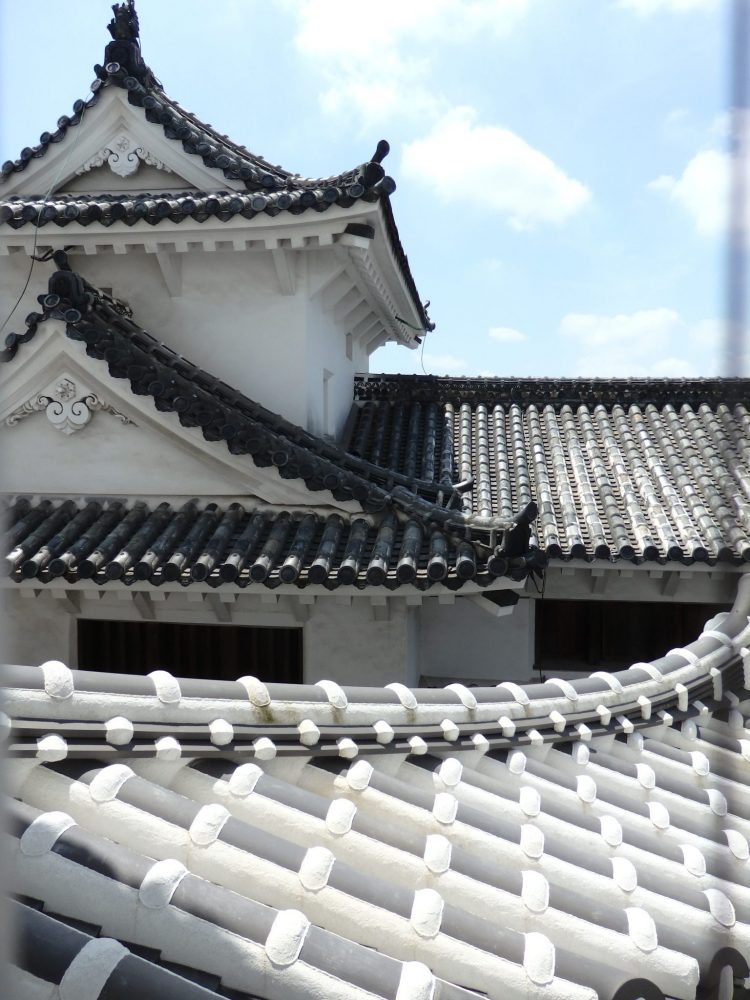 roof detail on Himeji Castle: behind is a grey roof, with rounded tiles in lines, the gables of the roof curved. Nearby a small part of a white roof is visible, again with curved tiles, but also with regular decorative crosspieces.