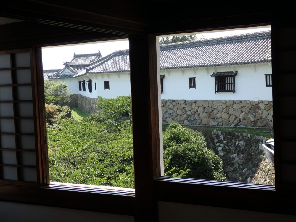 View of the West Bailey from Himeji Castle. A window frames the view, and the baily is only one story tall, white sided, but resting on a brown stone foundation.