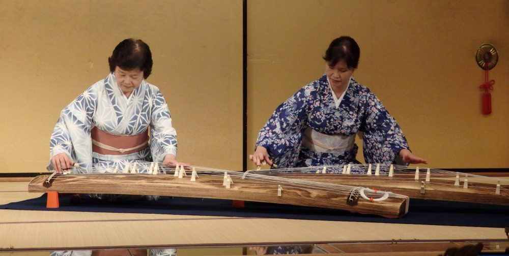 two women in kimonos sit on the floor, each playing a large harp that sits on the floor in front of them, in Gion Corner