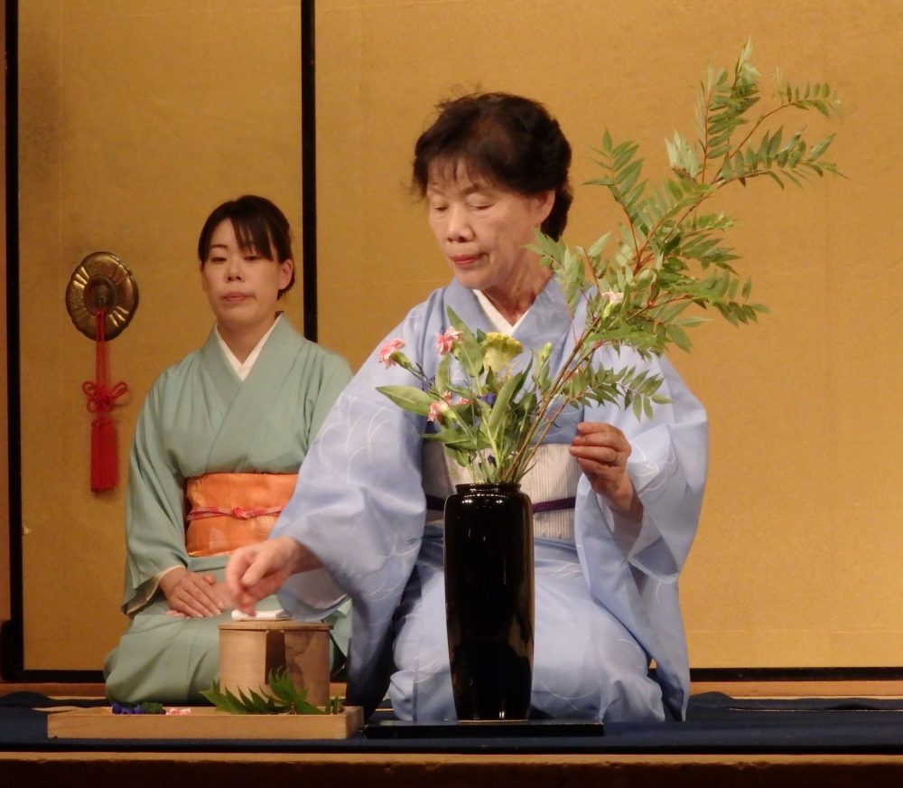 a woman in kimono kneels, arranging leaves and flowers in a vase in front of her on the floor, in Gion Corner