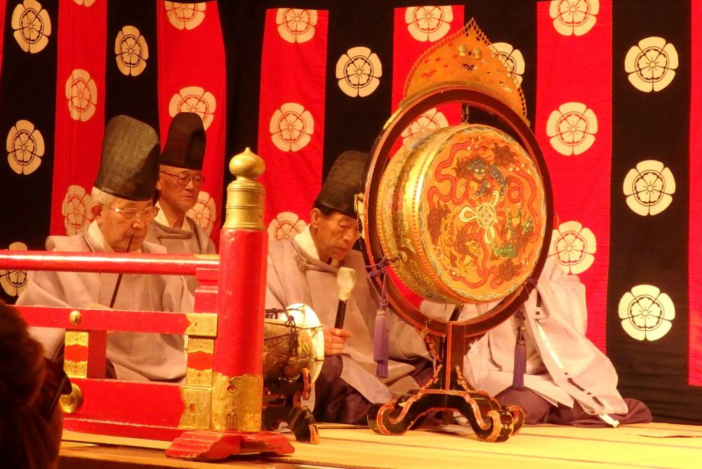 a group of men sit on the floor at the side of the stage. One bangs a large gone, while the others play other instruments, including flutes. Gion Corner