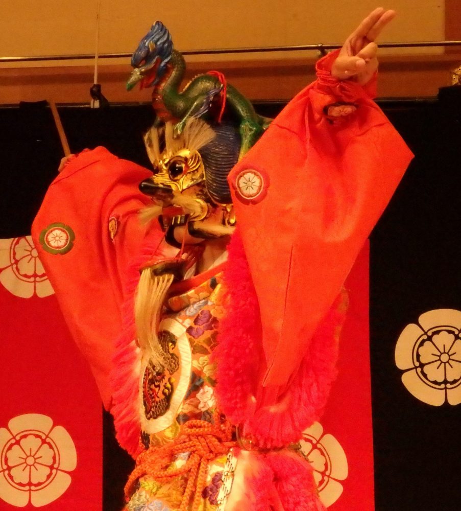 The dancer wears an elaborate, mostly red and gold, costume, and his face is covered with a fierce mask, in Gion Corner