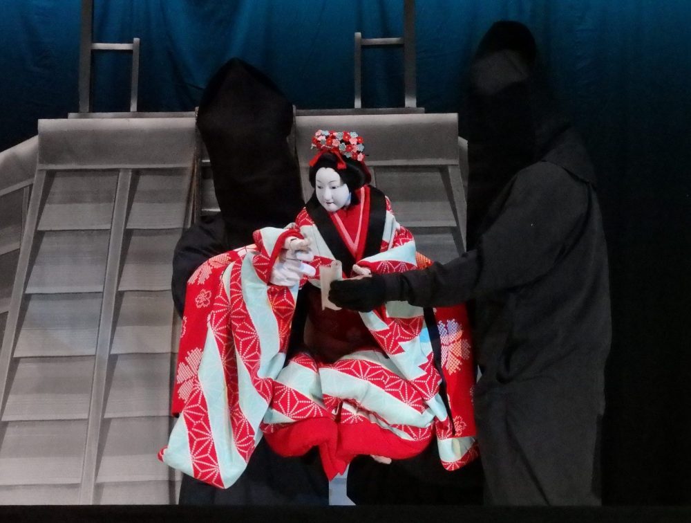 The puppet, dressed in an ornate, flowing, kimono, is carried by two puppeteers: men dressed entirely in black, including hoods over their faces. Gion Corner