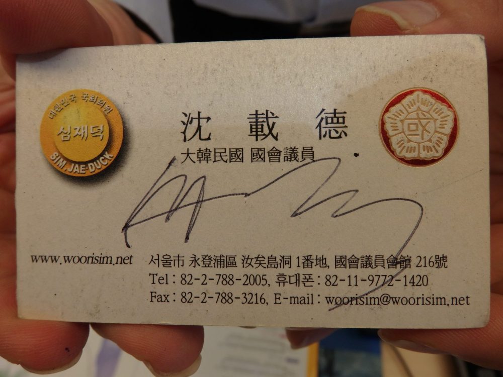 The business card is pretty typical: beige background with the info in black, mostly in Korean, except that Mr. Toilet House's signature is scrawled across the middle. There are two crests or logos, on on either side. The left-hand one is yellow and, besides the Korean lettering also says Sim Jae Duck. The right-hand one is some symbols inside a 5-sided crest. 