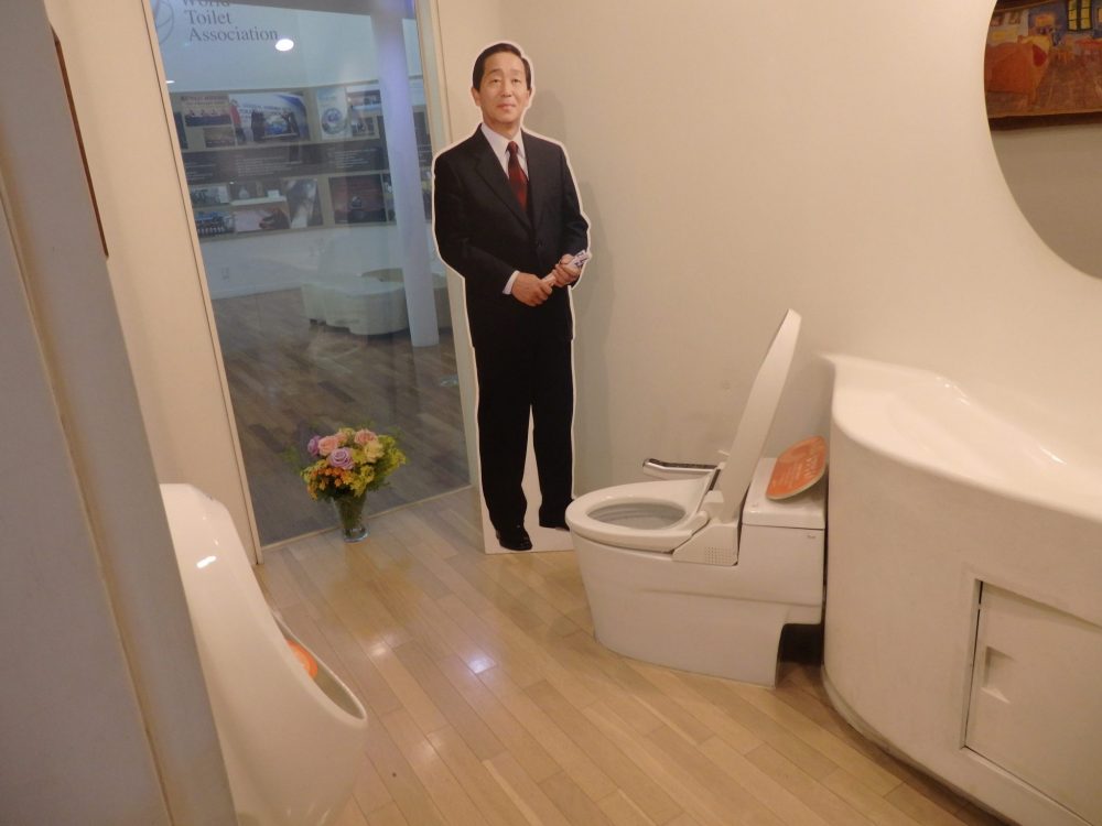 The bathroom has a window in the background looking out on what is now exhbit space. A vase of flowers stands on the floor in front of it. The toilet stands on the diagonal, facing the window at a slant. Next to it is a life-size standing image of Mr. Toilet House in a suit and tie. A mirror is partly visible on the right-hand wall and a urinal on the left-hand wall.